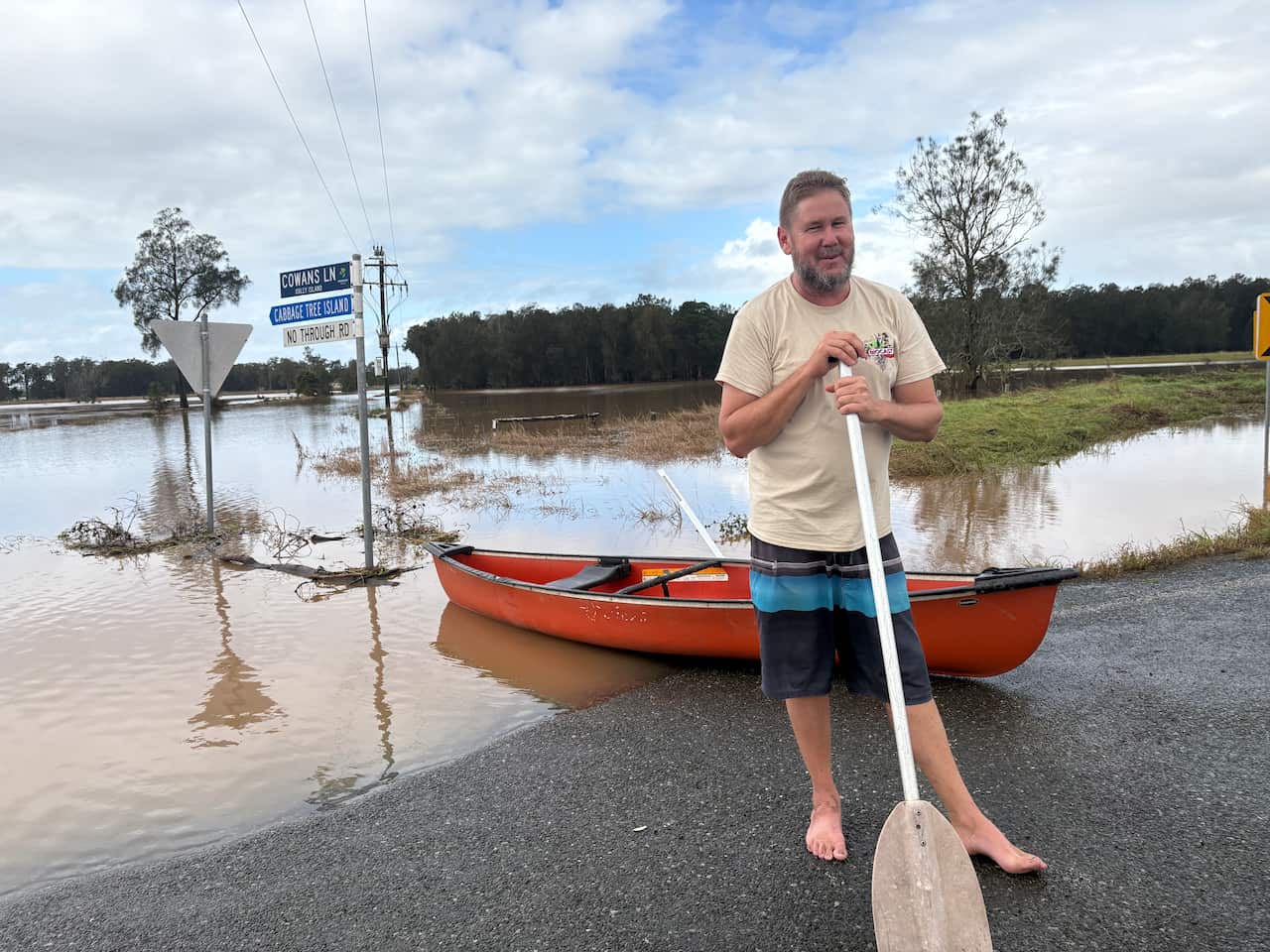 A middle aged man wearing yellow shirt and black and blue board shorts, standing barefoot on a road holding a canoe paddle, with an orange canoe sitting behind him in floodwater over the road.
