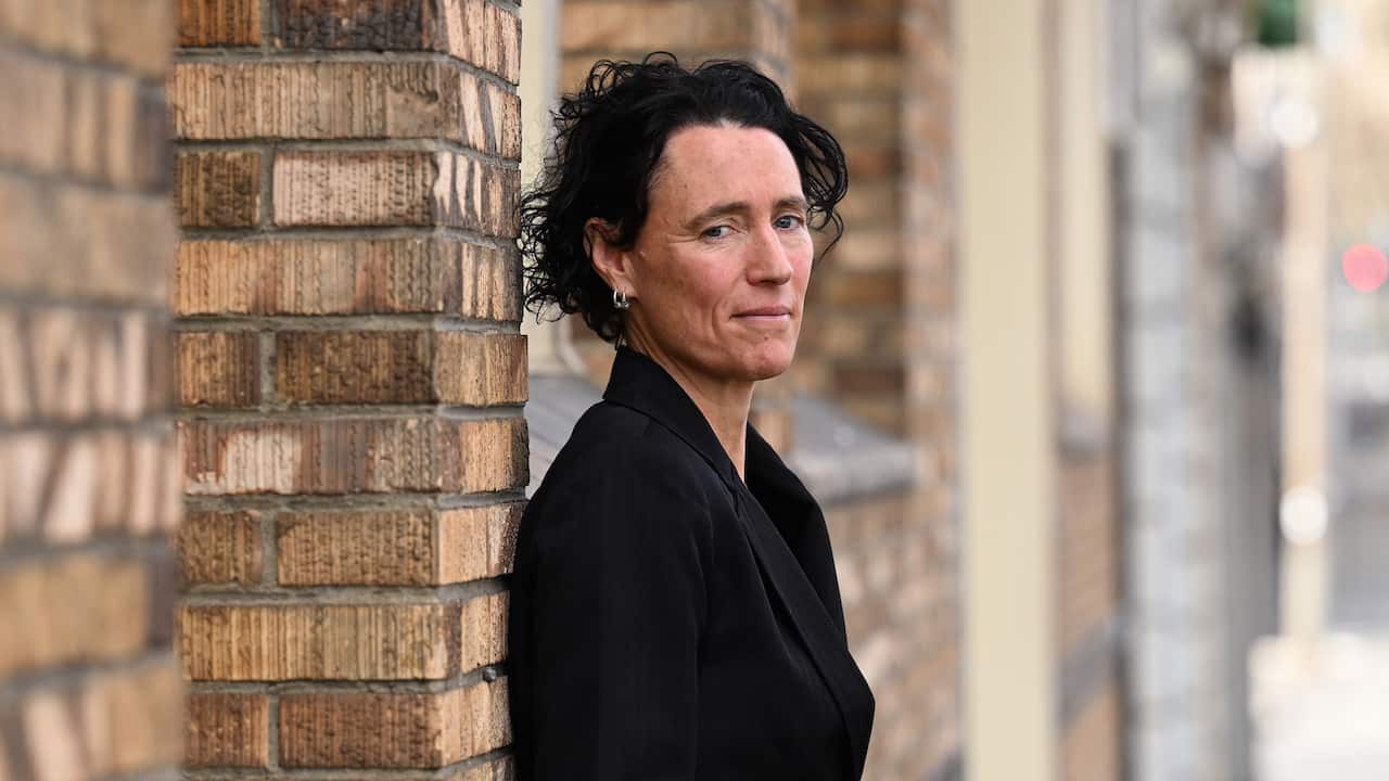 A woman with short black hair, wearing a black blazer, standing against a brick wall.