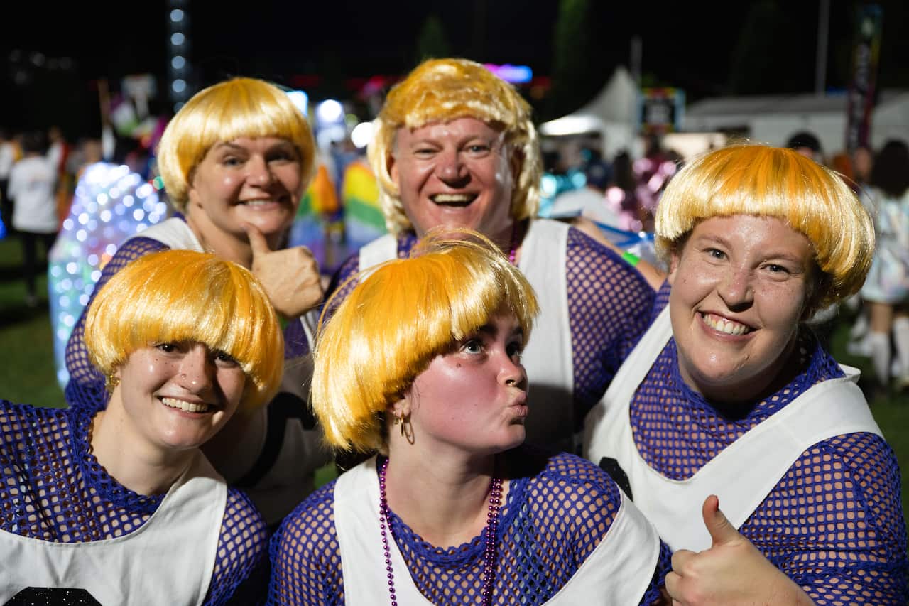 A group of smiling people wearing blonde wigs and netball jerseys hold thumbs up to the camera.