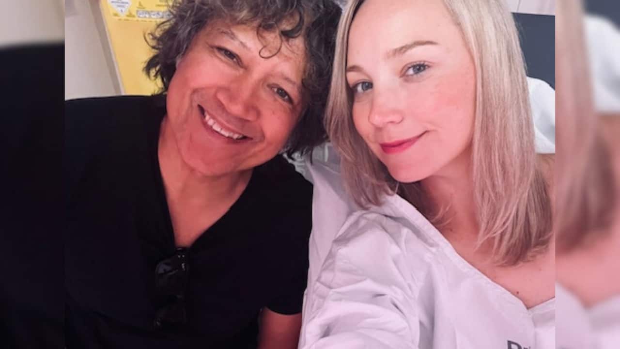 a blonde woman in a hospital gown and hospital bed smiles with a man in a black t shirt with curly hair