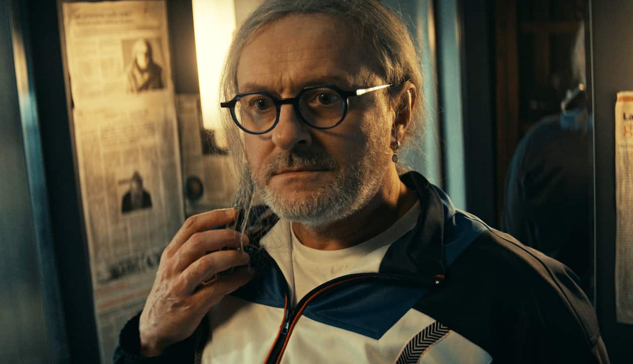 A man in glasses with white hair and a short beard stares ahead, with one hand raised towards his face. The wall behind him is decorated with pages from a newspaper. 