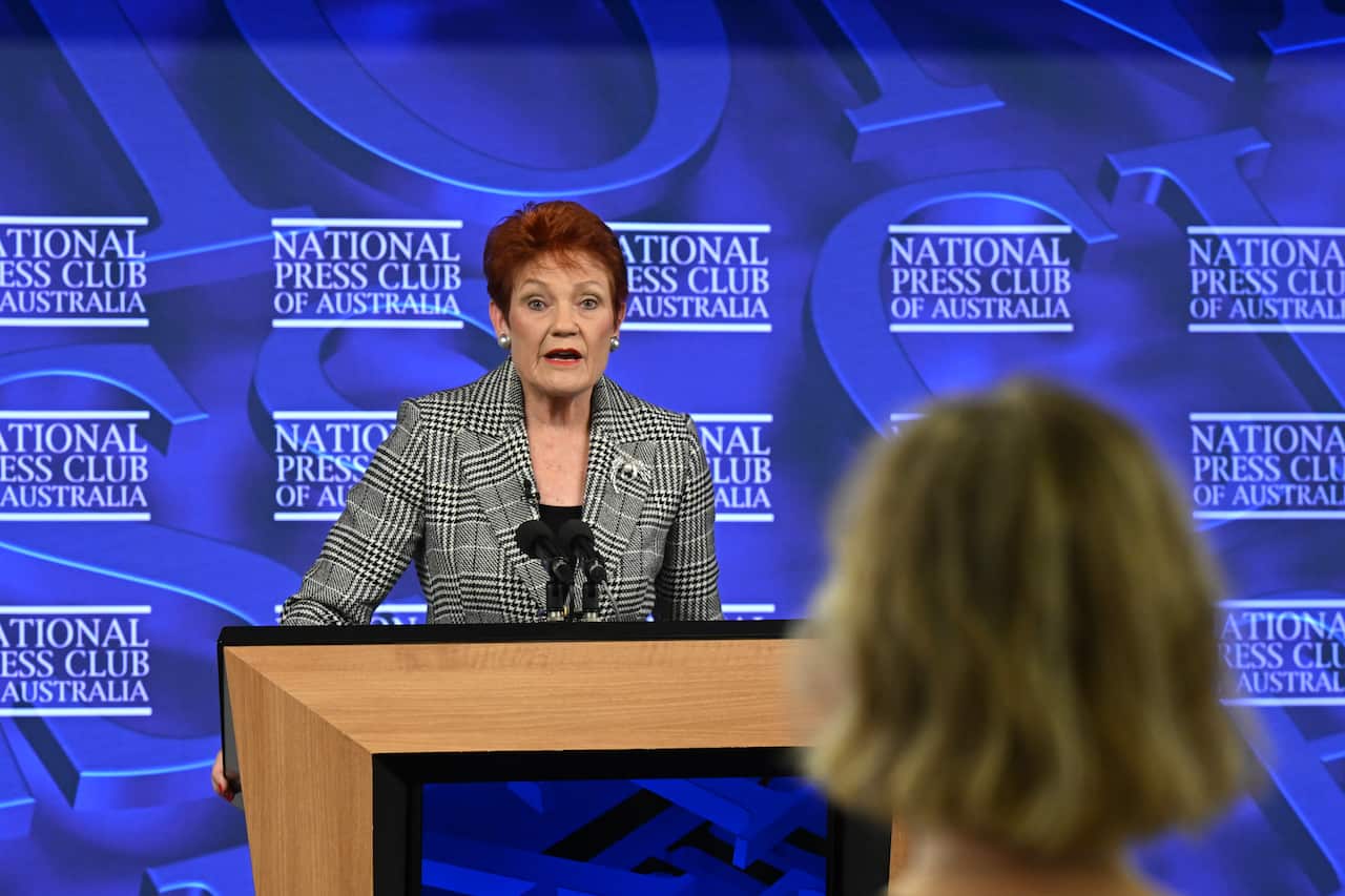 Hanson speaks from a podium with the back of a journalist's head visible in foreground