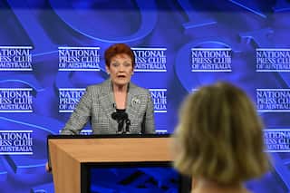 Hanson speaks from a podium with the back of a journalist's head visible in foreground