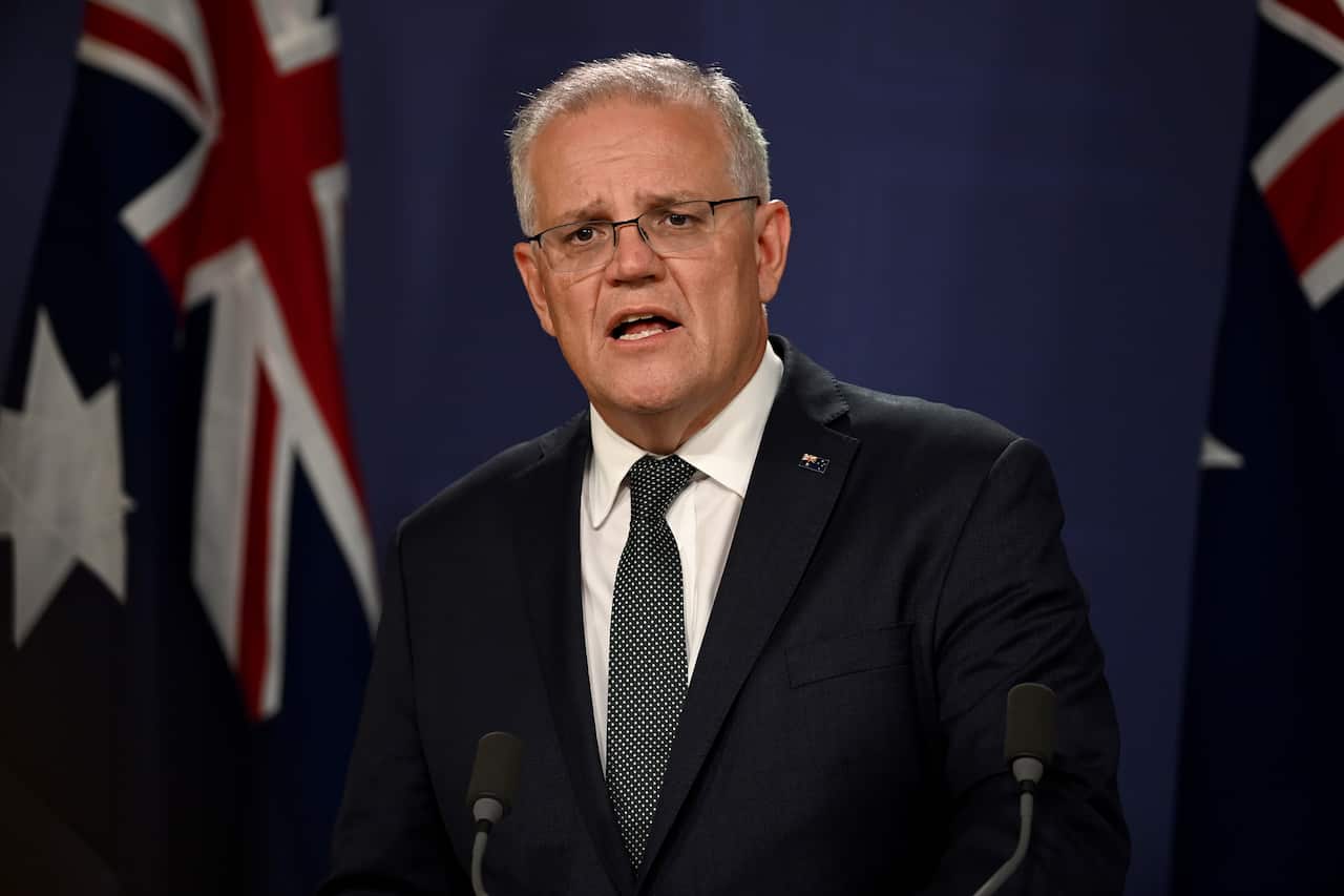 Scott Morrison stands in front of the Australian flag, behind a microphone perched on a podium. He is wearing a black suit and black tie, as he speaks