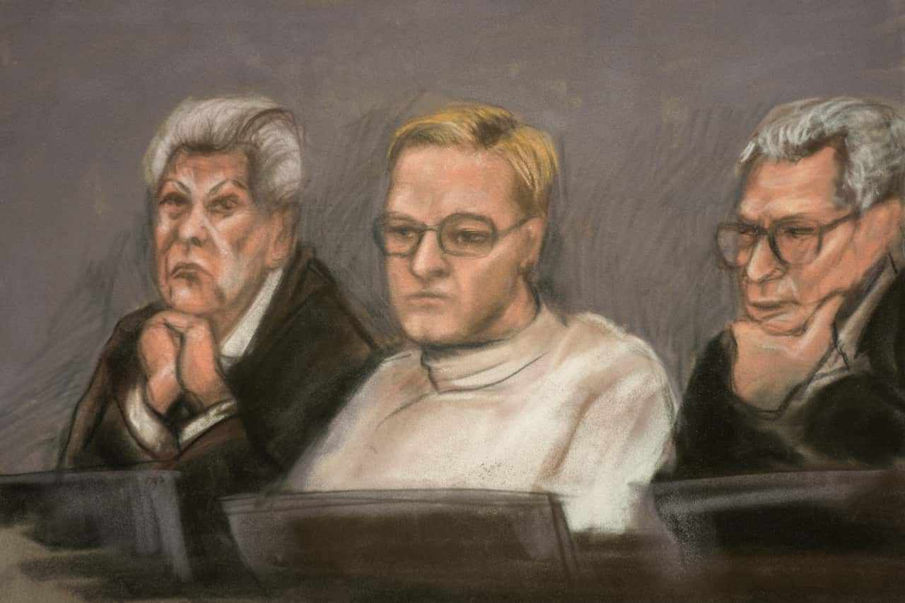 A court sketch of a man with blonde hair, wearing glasses and a white top. Two lawyers wearing suits and serious expressions sit next to him.