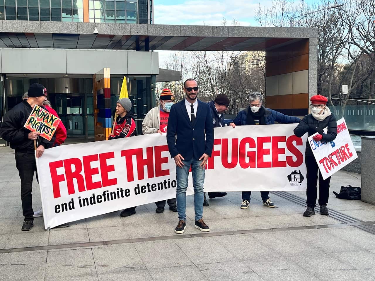 Moz stands in front of a big banner carried by multiple people with the words "free the refugees - end indefinite detention'