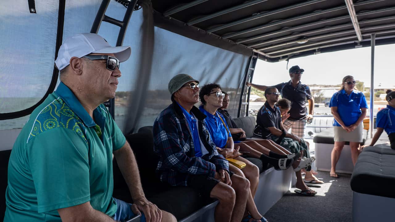 Passengers aboard a medium-sized fishing vessel sit listening to someone out of shot talking.