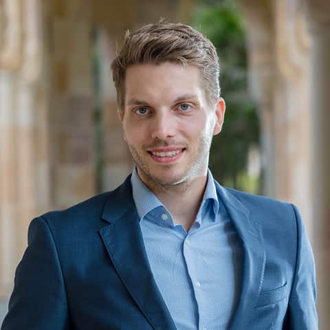 A man in a blue suit smiles into the camera, behind him is an out-of-focus sandstone building