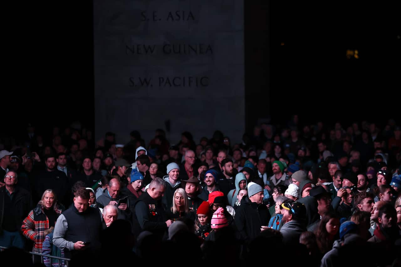 A crowd of people stand in the dark, a large marble structure is semi-illuminated behind them.