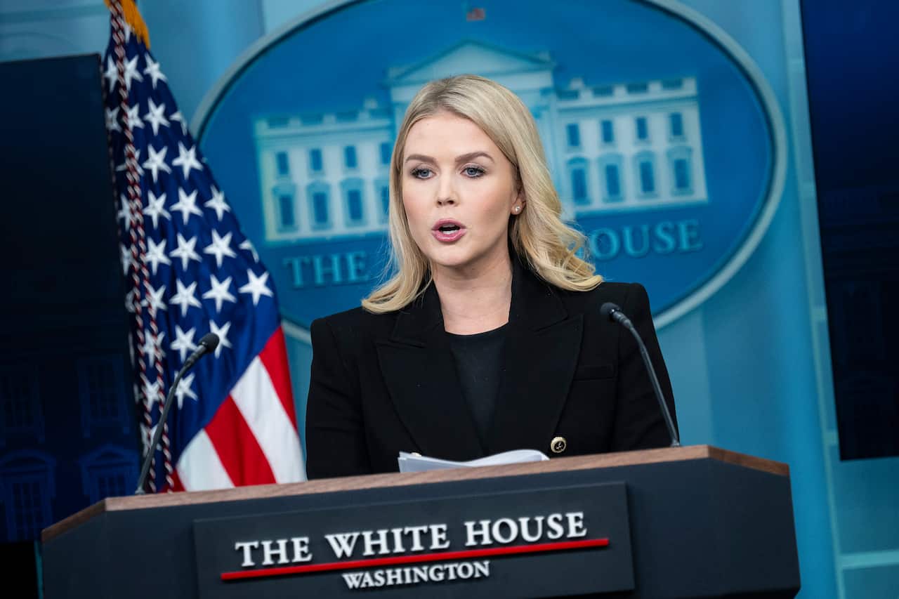 A woman in a dark suit stands in front of a podium with a sign that reads "The White House" 