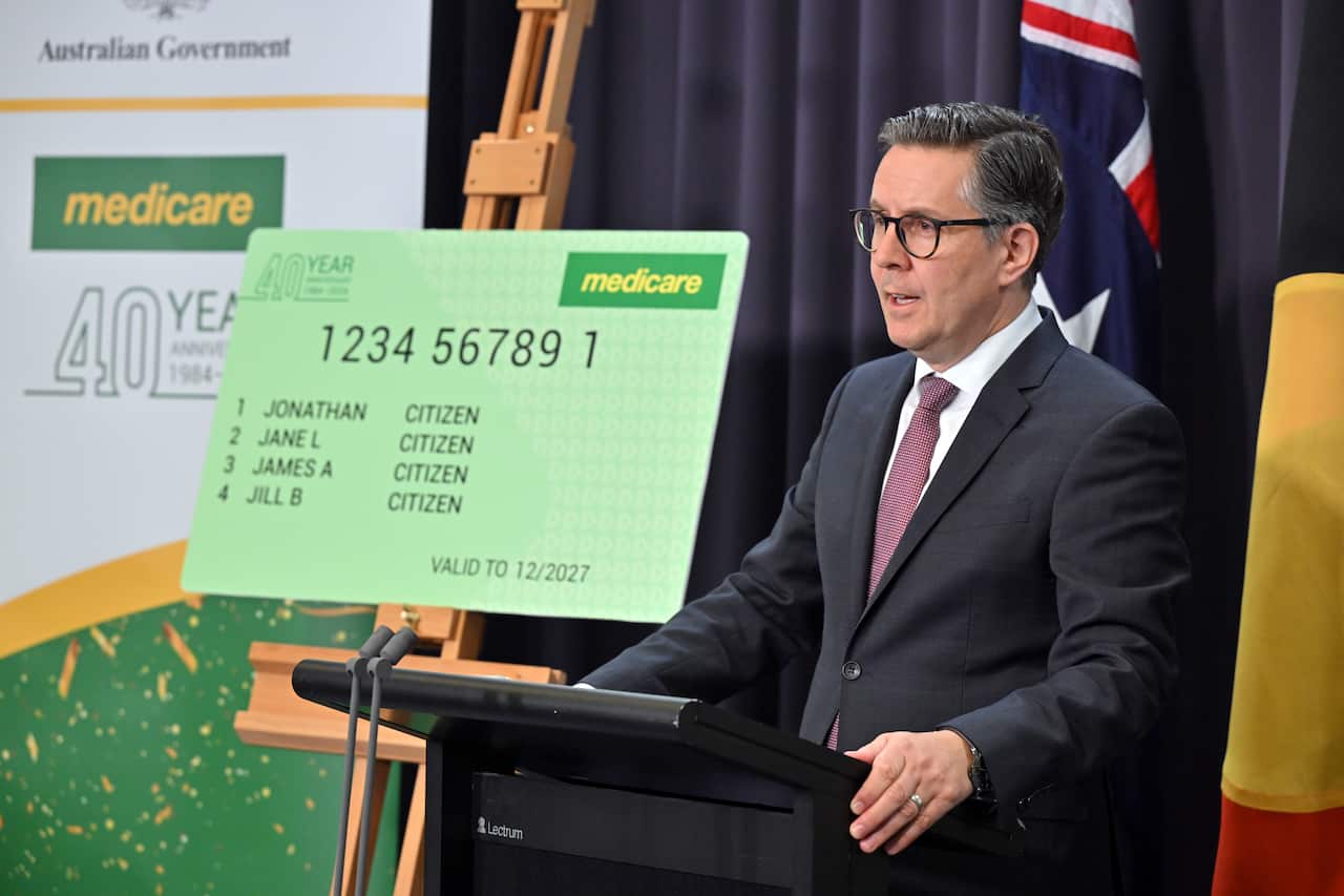 A man wearing an official suit and a tie is standing in front of the Australian flags, with a large Medicare card in the background.