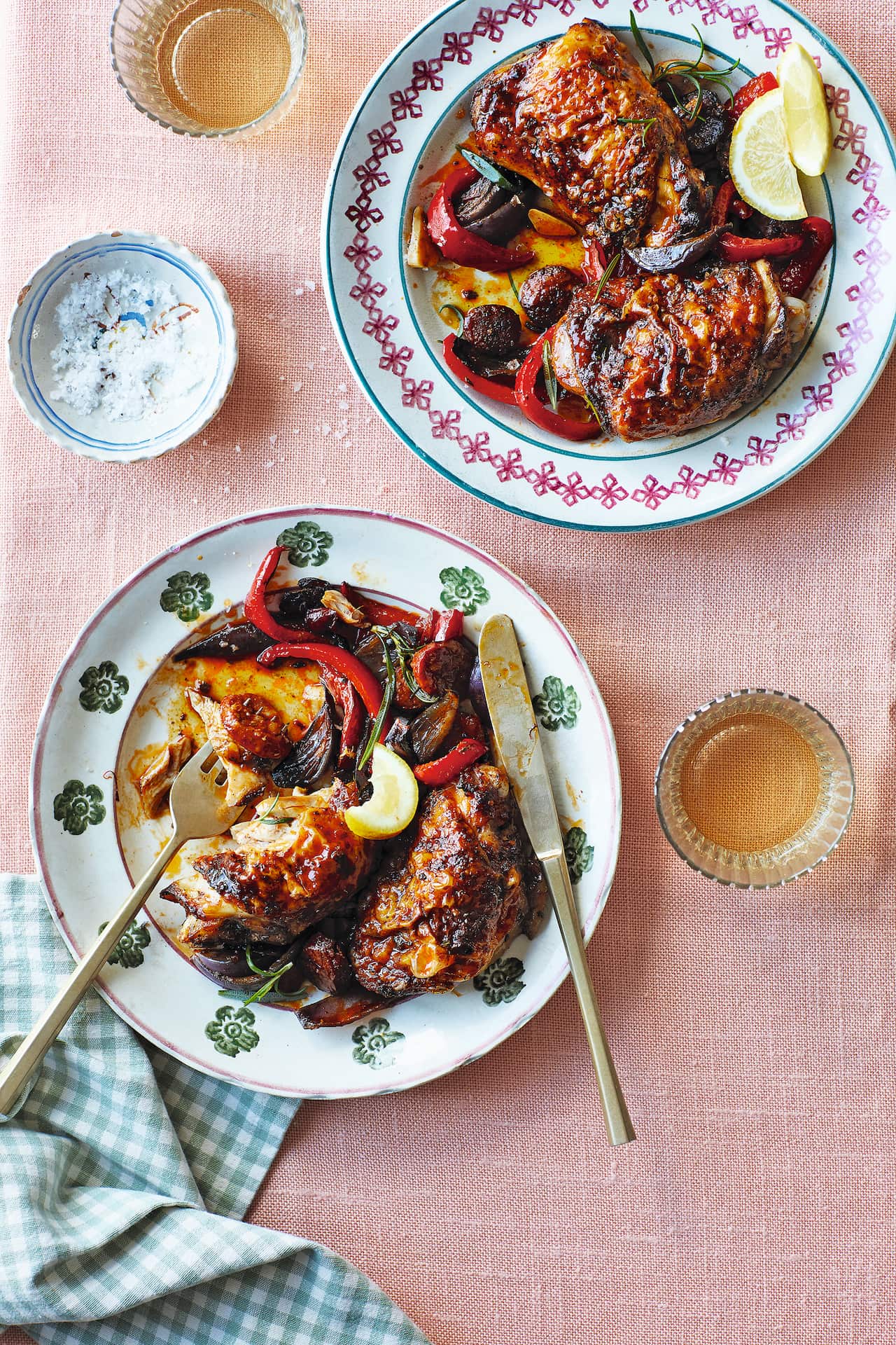 Two plates, holding serves of a chicken and capsicum dish, sit on a pale pink tablecloth. A folded napkin sits beside one plate. 