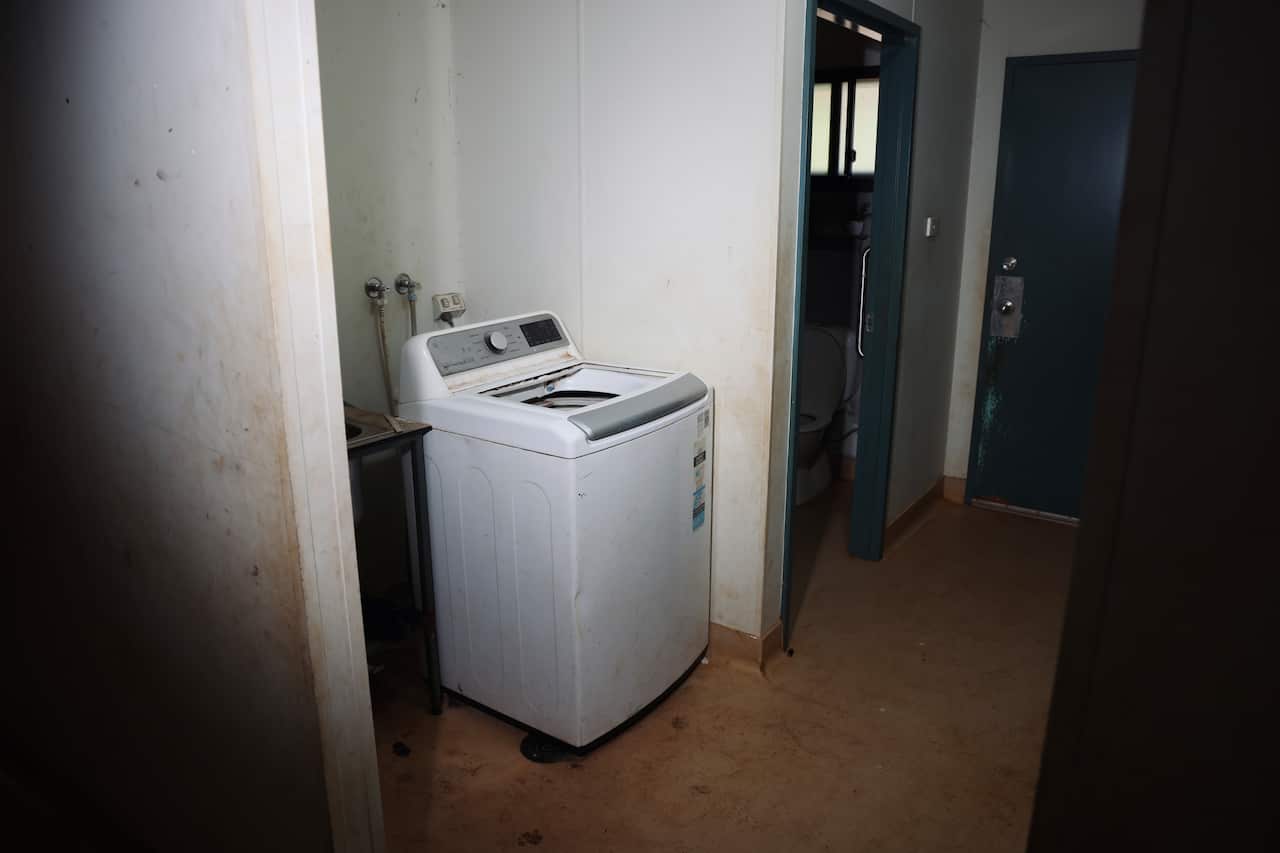 A top loading washing machine sits in a dark room, next to a toilet.