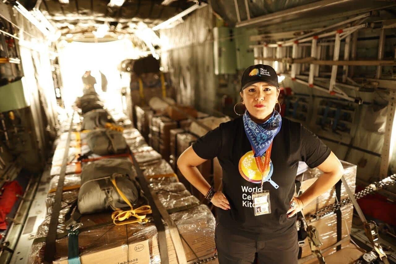 A woman with her arms on her hips, wearing a t-shirt and hat with the words 'World Certified Kitchen' on it. She's standing in front of aid packages. 