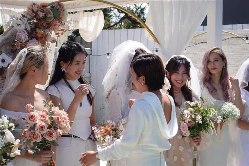Women wearing wedding dresses smile as they pose for a photo.