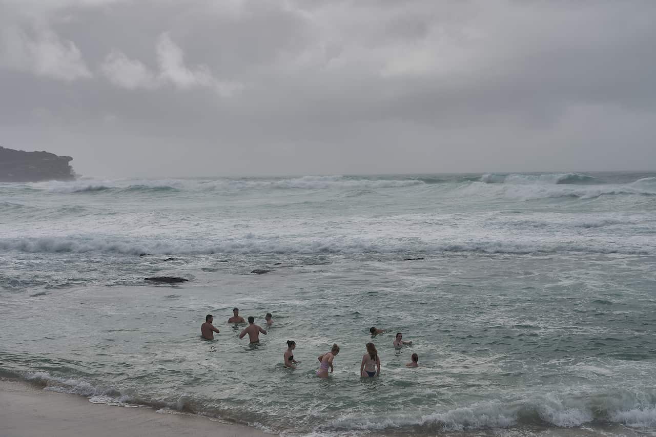 Around 10 swimmers in the shallow waters of a beach while beyond them wild surf rages.
