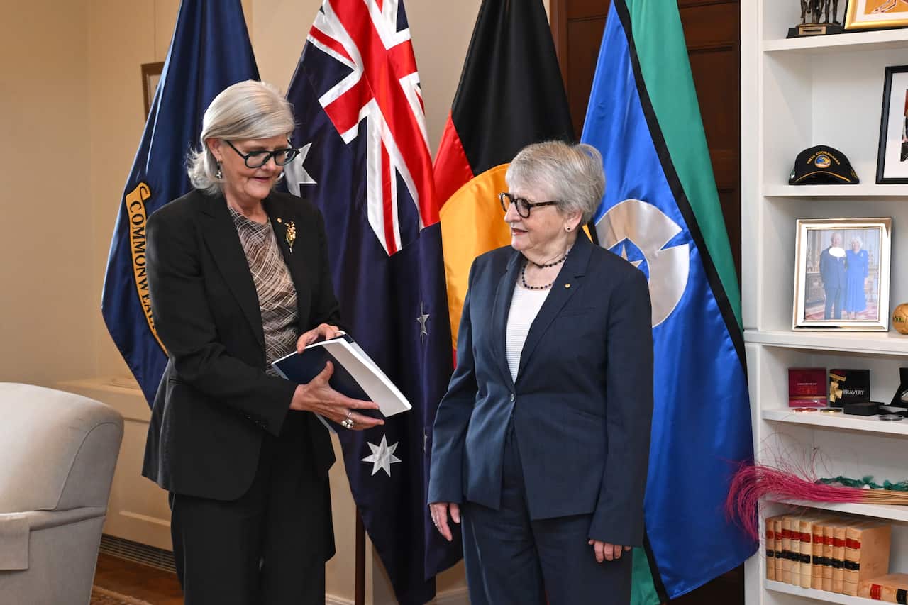 Two women standing in a room together. One is holding a folder with documents. There are Australian and Indigenous flags behind them.