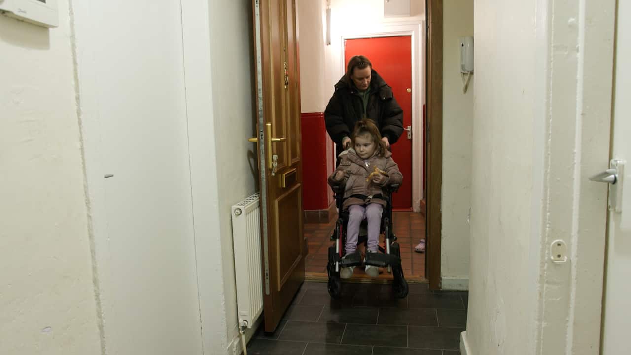 A woman in a black puffer jacket pushes a young girl in a wheelchair through the door of an apartment.