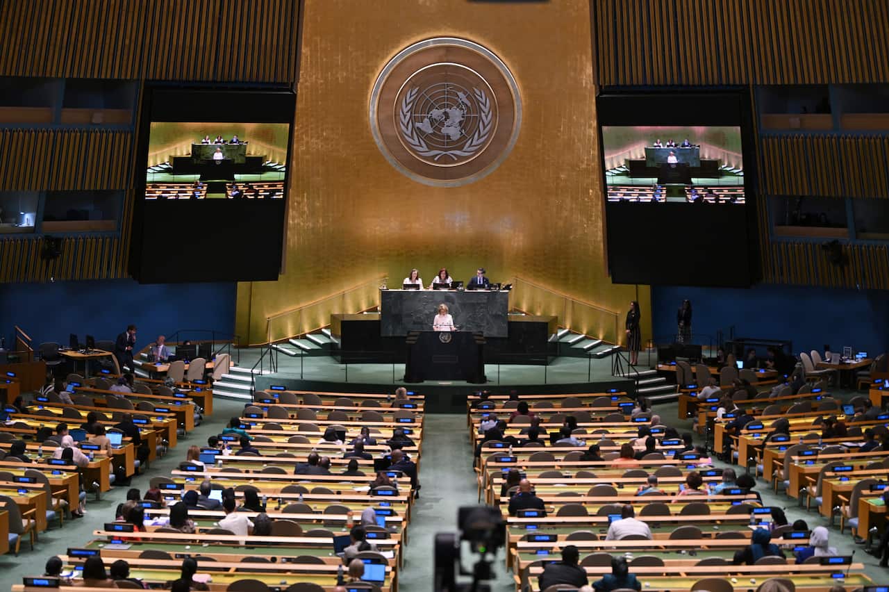 A woman speaks at a podium in front of a large auditorium filled with long tables and chairs. 