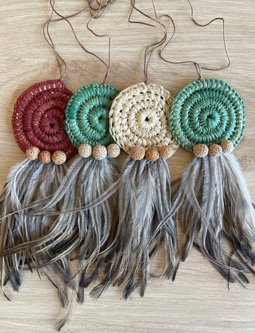 Four necklaces laid side by side, each consisting of a circular woven section connected to hanging grey feathers. From left to right, the colour of each woven section is: maroon, teal, tan, and teal again. 