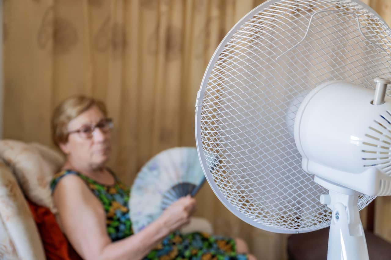 An elderly woman sits on a sofa, holding a hand fan, with an electric fan blowing in front of her.