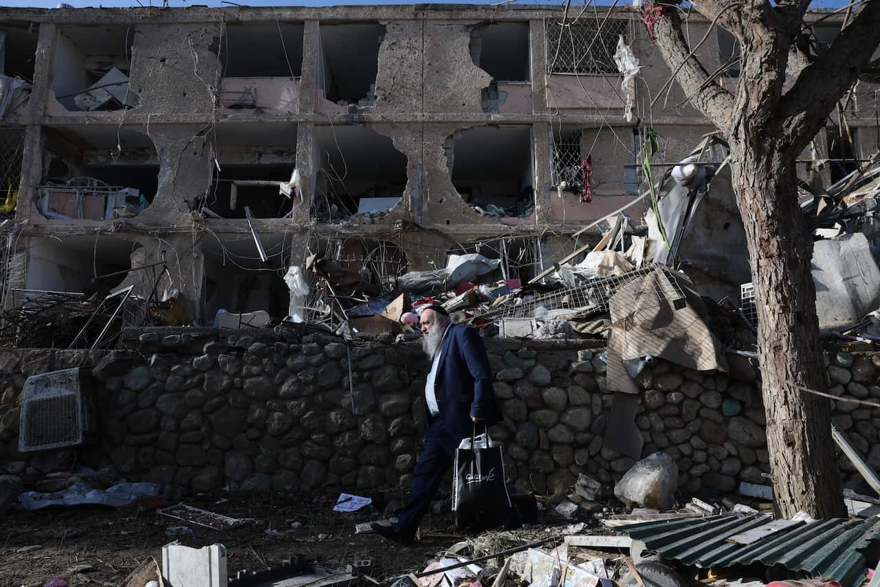 An ultra-Orthodox Jewish man carrying a shopping bag walks past a heavily damaged multi-story residential building surrounded by rubble and debris.