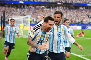 A football player wearing a light blue and white jersey is elated after scoring a goal. His teammate in a light blue and white jersey is pulling his shirt as he also celebrates with him. Two other teammates are seen running behind them, smiling.