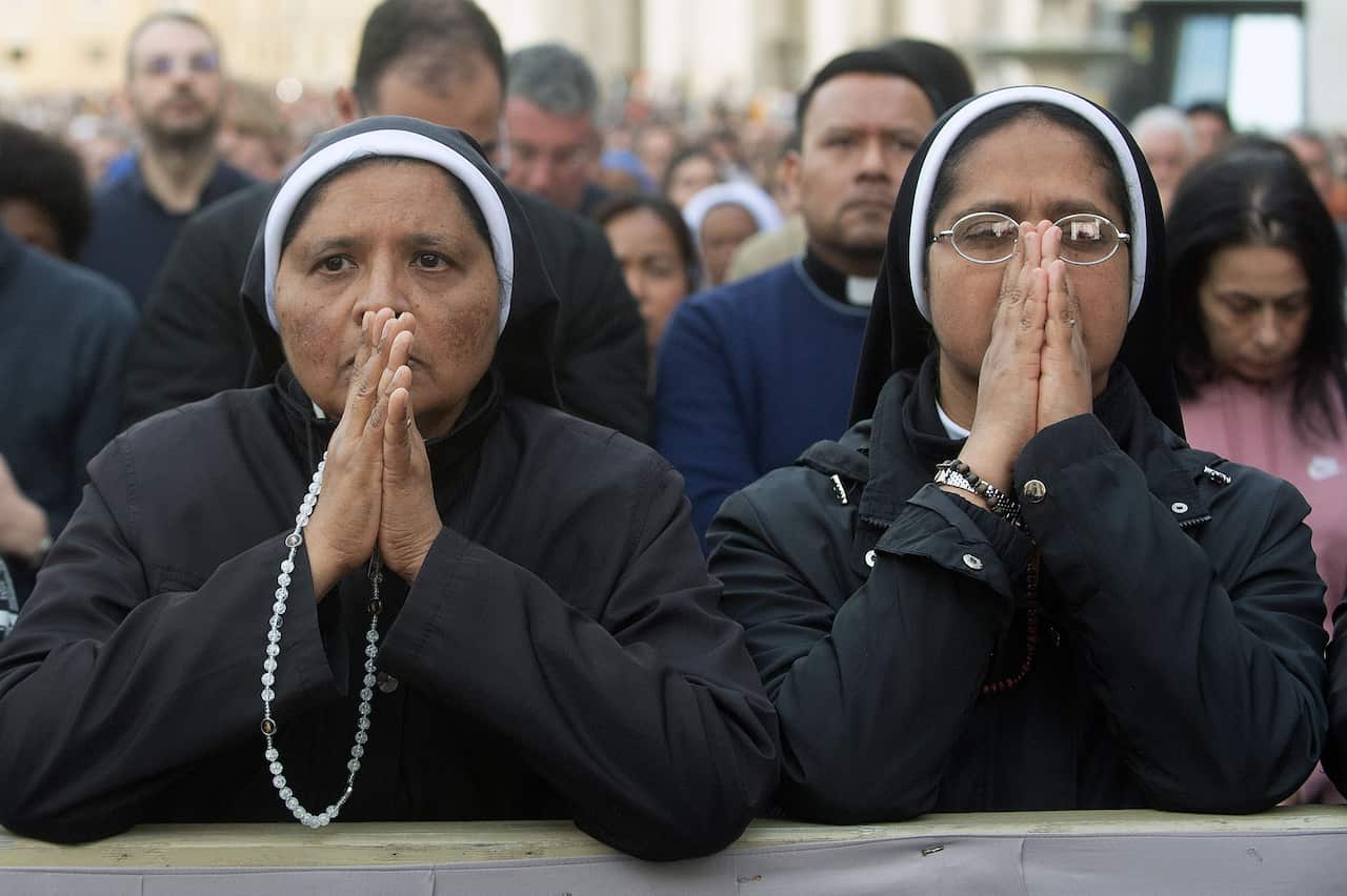 Two nuns dressed in black robes have their hands folded in front, with a crowd visible behind them.