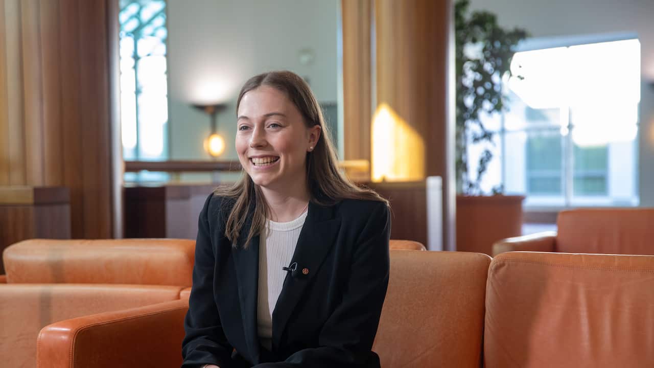 A young woman in a white tshirt and black blazer smiling as she sits on an orange couch.