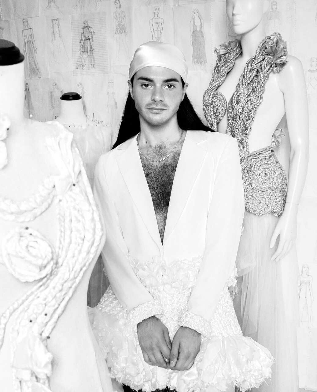 A black and white photo of a person with long hair wearing a low-cut dress and head scarf, surrounded by mannequins with their designs on them