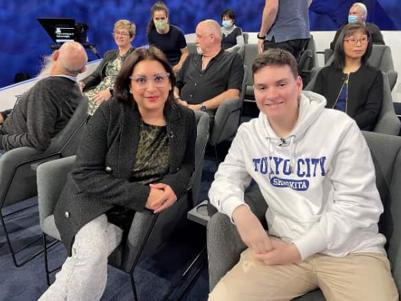 A screenshot of a mother and son seated together. There are other people seated behind them.