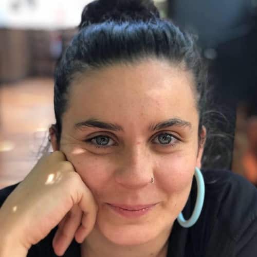 A woman with a bun and light blue hoop earrings smiles, her hand on her cheek.
