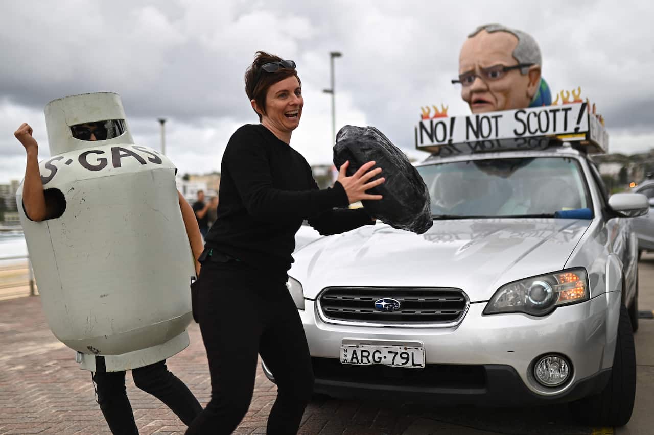 A woman wearing a gas canister costume dances with another woman holding a prop resembling a piece of coal in front of a vehicle mounted with a giant head of Australian Prime Minister Scott Morrison.