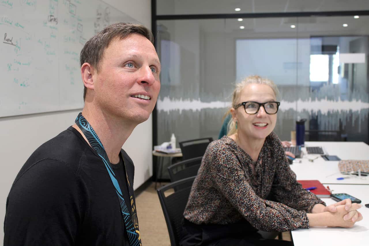 A man and a woman sit at a table in a meeting room, looking up as if watching a presentation. Scribbled words can be seen across a whiteboard behind them.