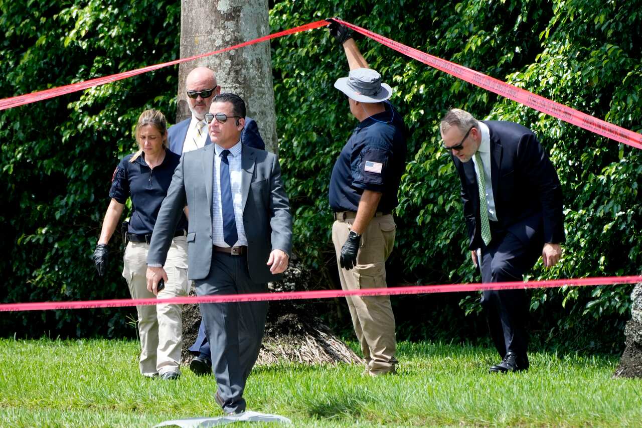 Four men and a woman on an area of grass near bushes. One man is holding up red police tape so another man can step under it
