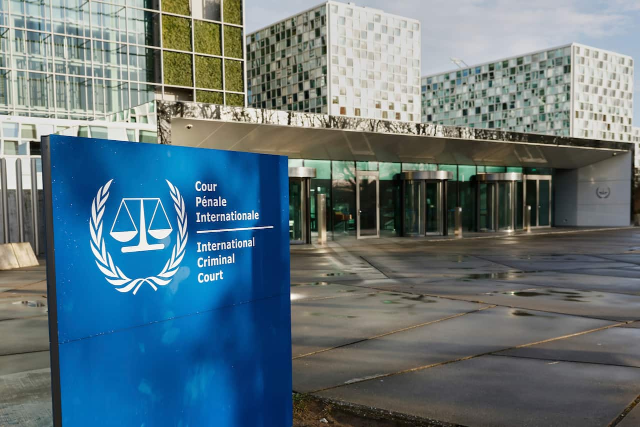 The official sign of the International Criminal Court is displayed outside its headquarters in The Hague. The sign is mounted on a wall or structure, with the modern ICC building and glass façade visible in the background.