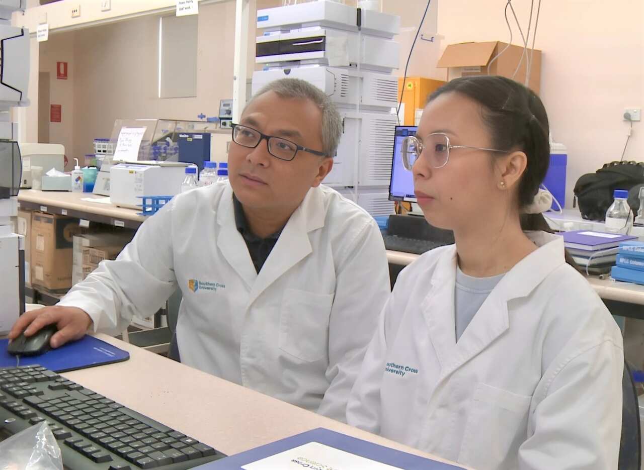 A man and a woman in white lab coats sitting next to each other look at a computer screen.