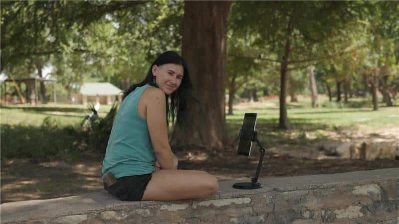 A person with dark brown hair sits on a low wall in a park. They are looking towards the camera and smiling slightly. In front of them is an ipad held by a tripod. 