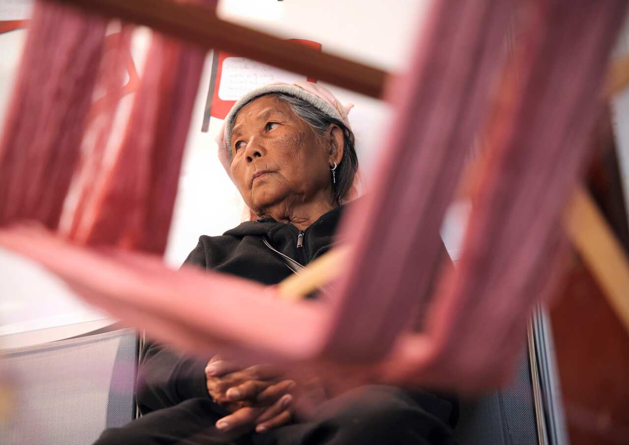 An older woman sits behind a wooden loom covered with pink thread.