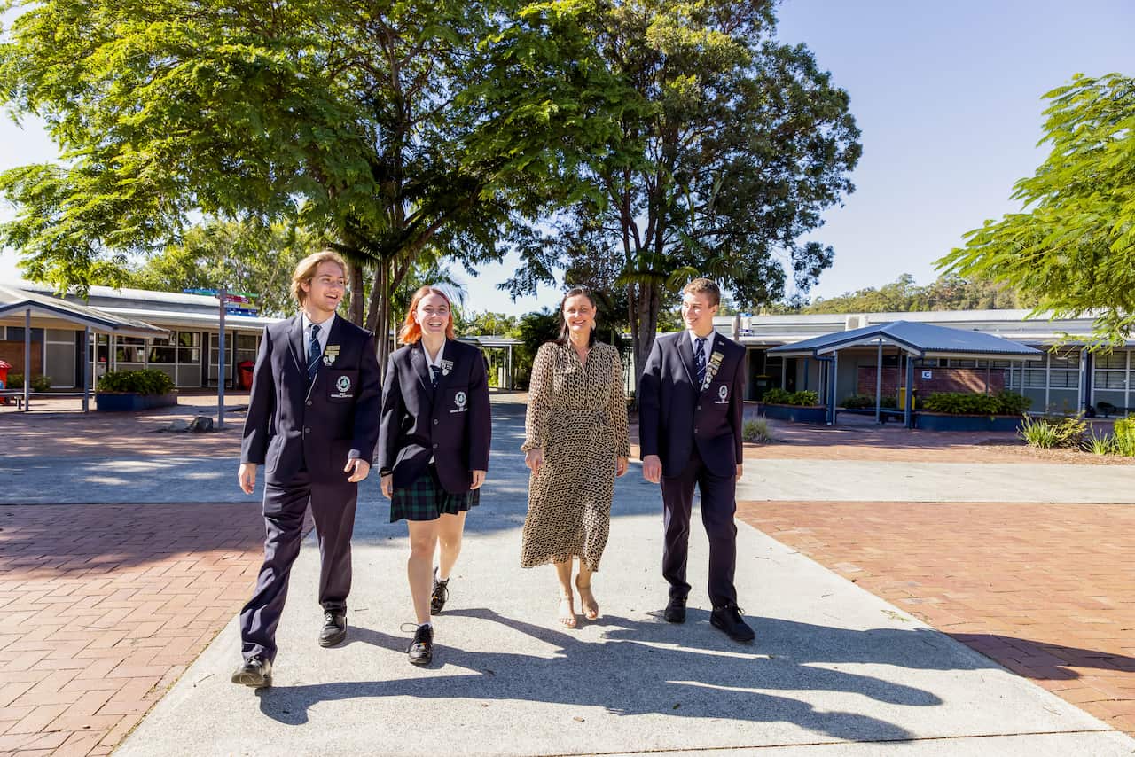 A woman walks with some high school students in the school grounds. It's a sunny day and they're all smiling.