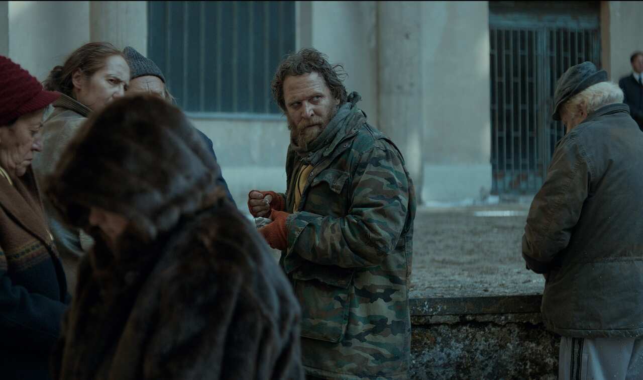 A group of people huddle on a street. A man in the middle looks warily over his shoulder. 