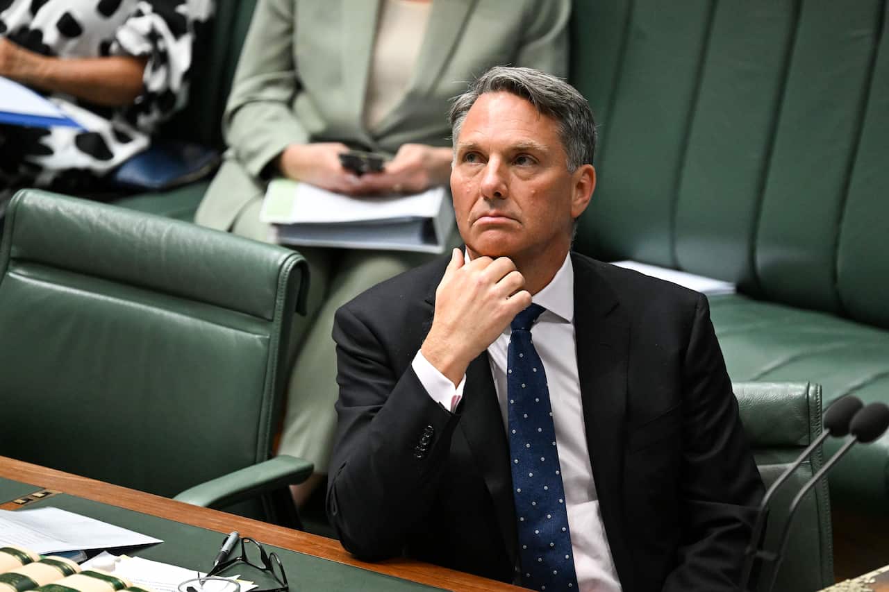 A man in a black suit and blue tie looks on with his hand on his chin.