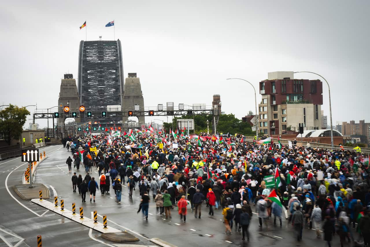 A large crowd of pro-Palestinian protesters marched across Sydney Harbour Bridge on a rainy day, carrying flags and signs.