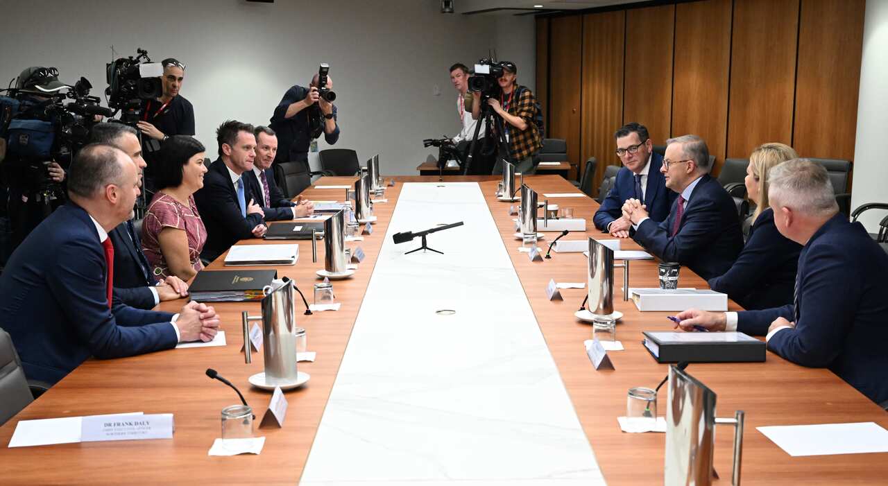 Group of people sit around a formal table in front of cameras.