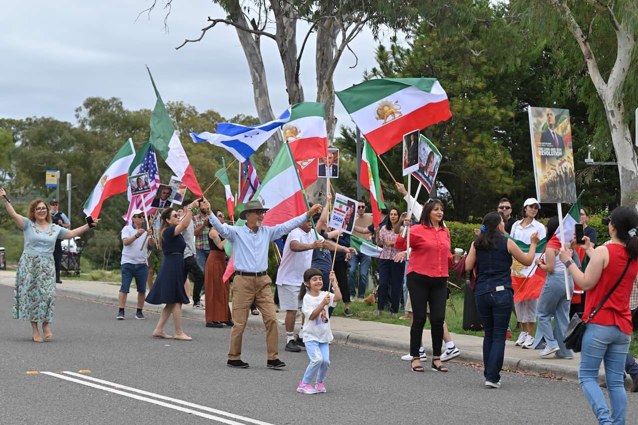 A group of demonstrators marches down a street carrying American flags and Iranian Lion and Sun flags alongside posters featuring Rezā Pahlavi.