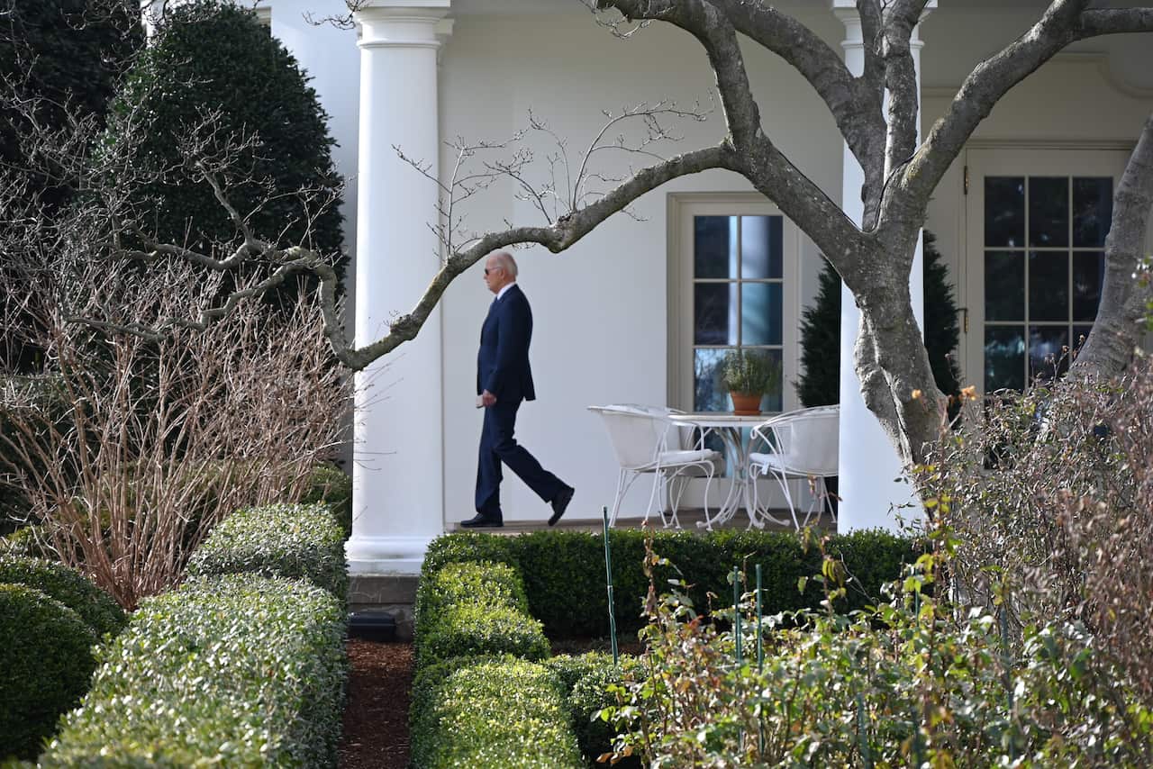 A man wearing a suit, tie and sunglasses walking along a verandah. 