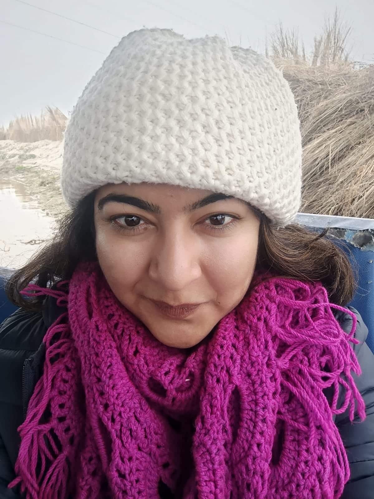 A selfie of a woman in a white woollen beanie and purple scarf, with the beach in the background. She has a neutral expression on her face.