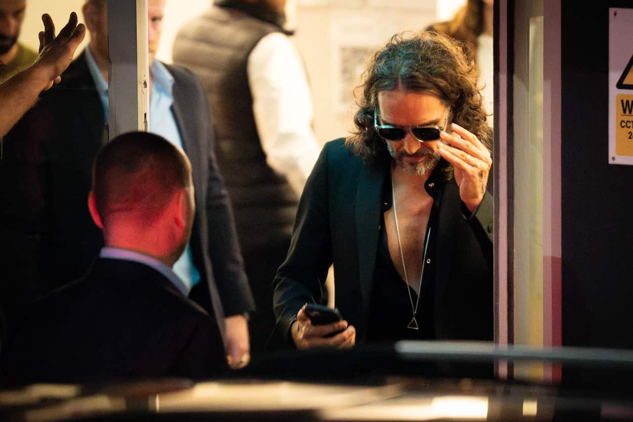 Hollywood actor Russell Brand dressed in black with his face down looking at his phone while attempting to take off his sunglasses.