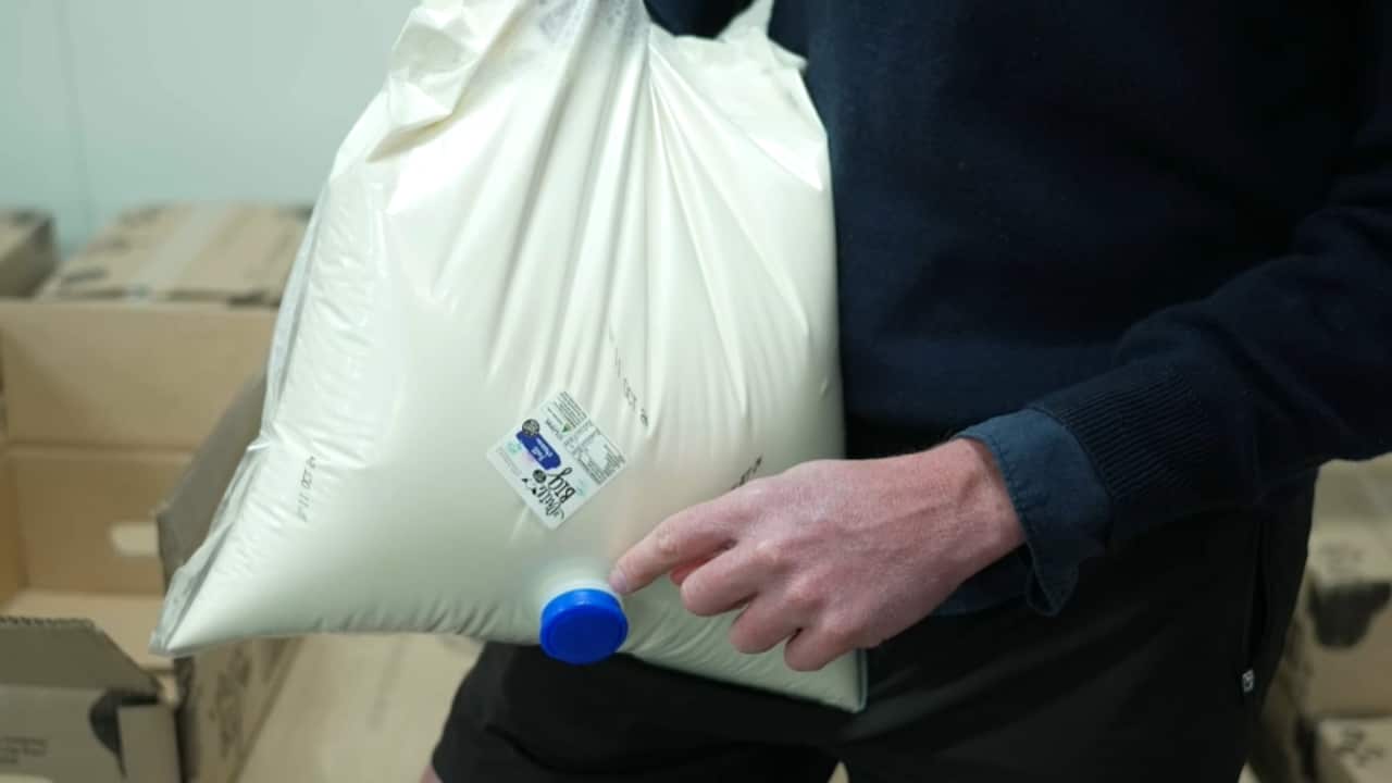 A white man holds a soft clear plastic bag filled with milk. It has a nozzle on it with a blue cap.