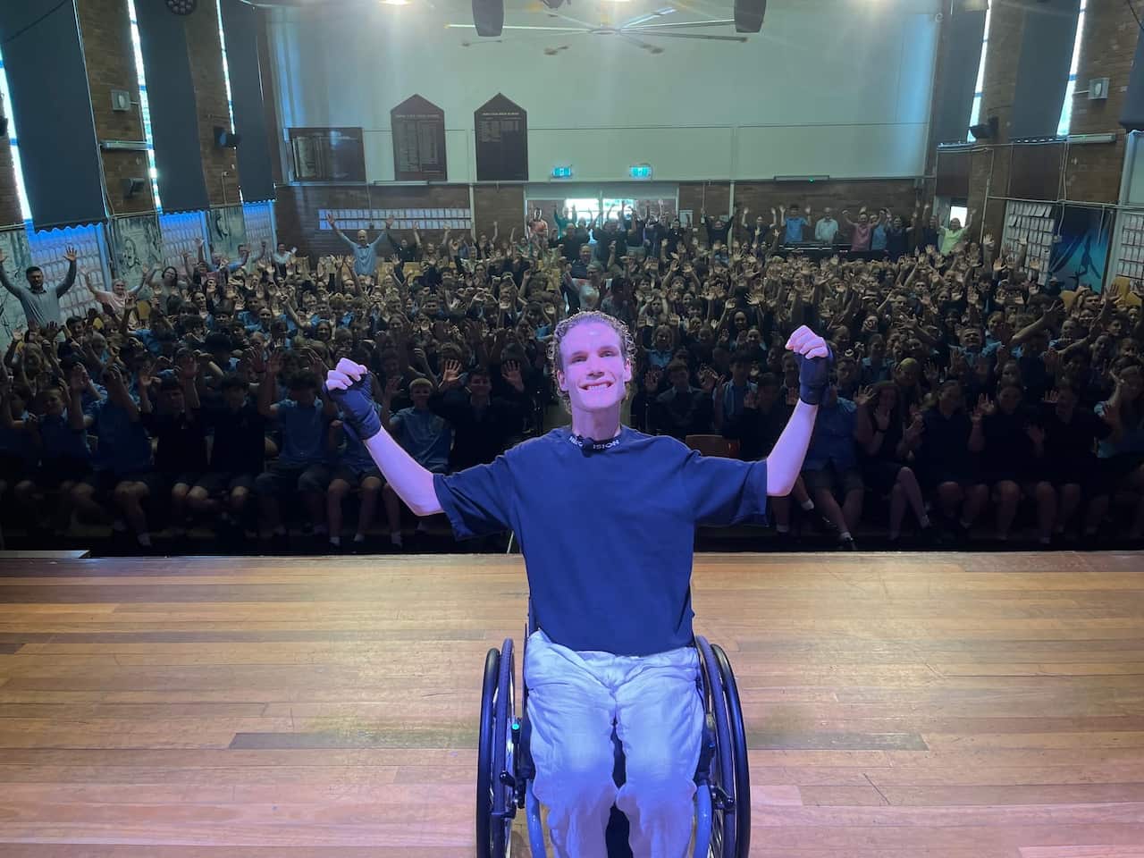 A young man in his wheelchair on a stage smiles with his arms in the air. Behind him, members of the audience also have their hands raised.